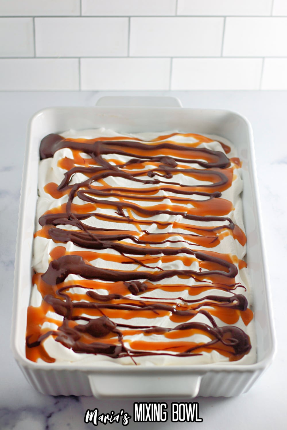 chocolate sauce and caramel sauce being poured on top of a whipped cream layer in a white casserole dish