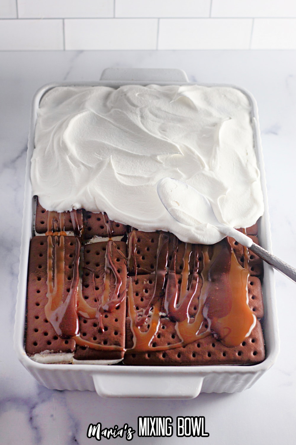 whipped topping being spread on top of a chocolate and caramel layer in a white baking dish