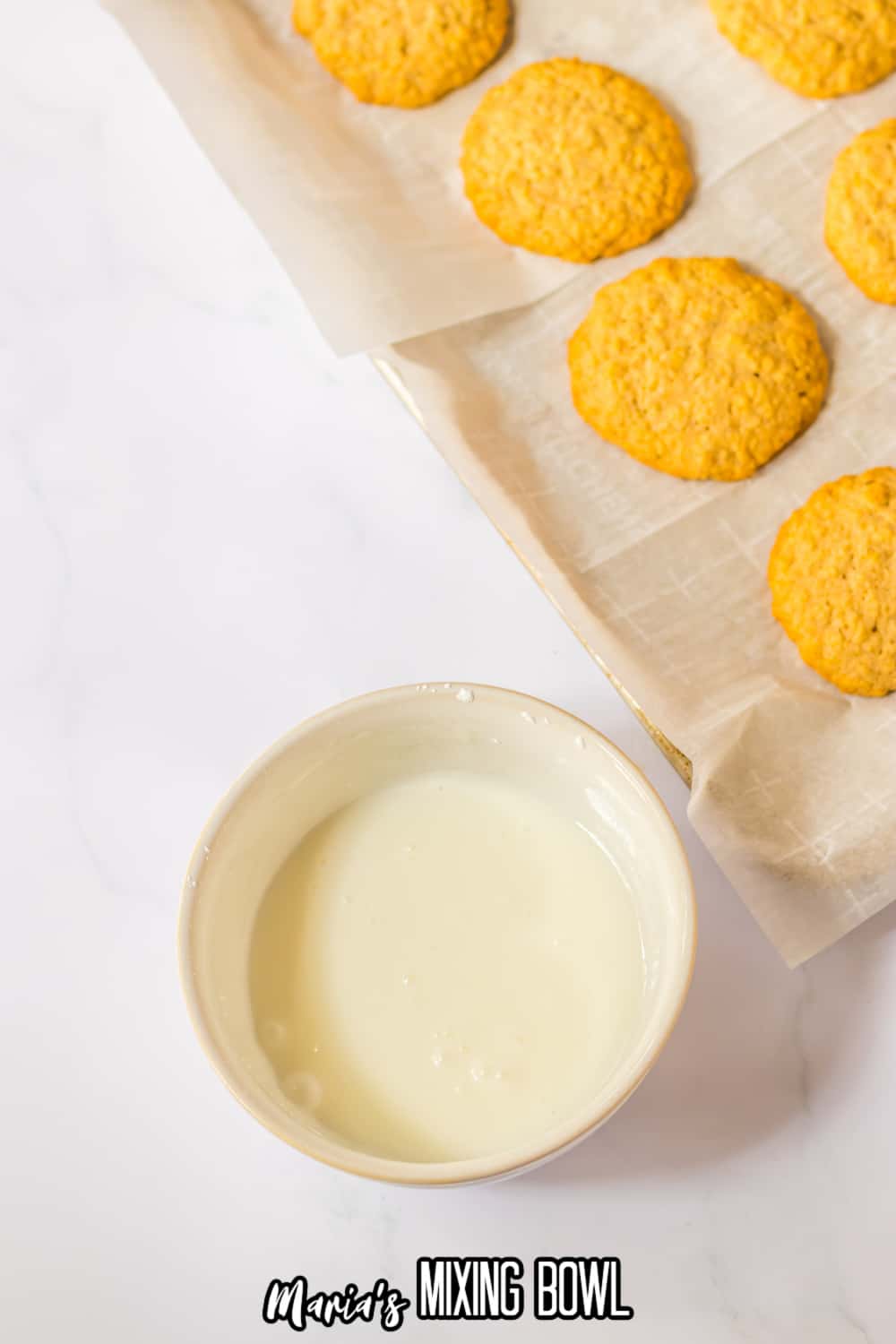 the icing for oatmeal cookies in a small white bowl with a tray of baked cookies behind it