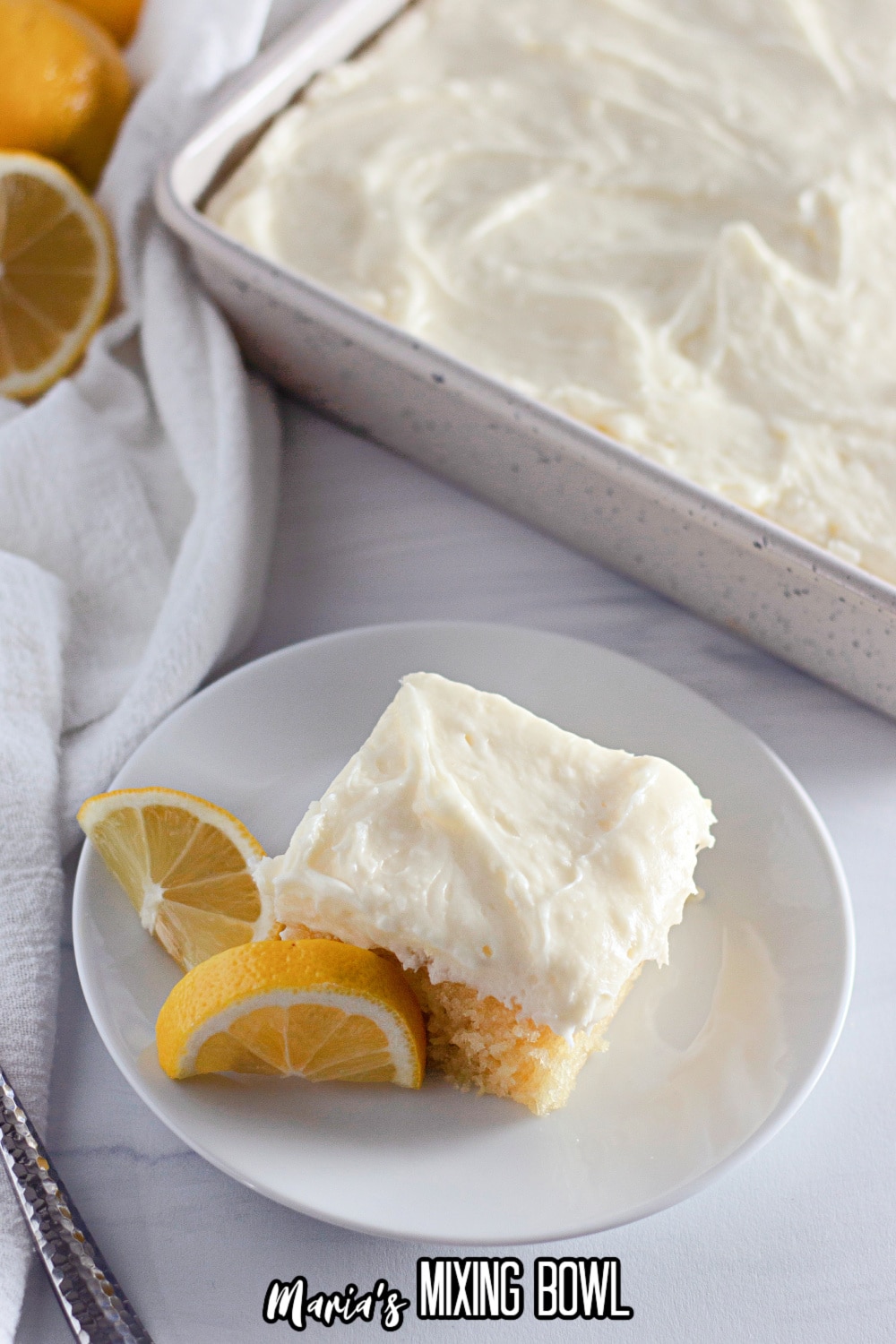 slice of lemon crazy cake frosting with cream cheese frosting on a white plate with lemon wedges