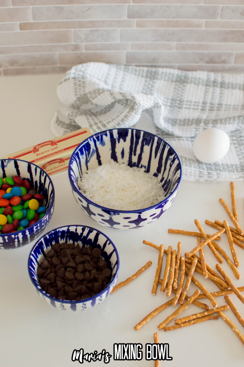 bowls of coconut, chocolate chips, m&M's butter, egg, and pretzels on a white backdrop