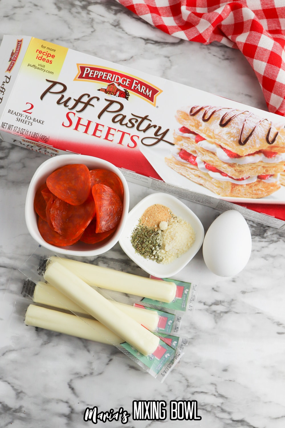 box of puff pastry, bowls of pepperoni, parmesan cheese, parsley, and garlic, an egg and string cheese on a marble background