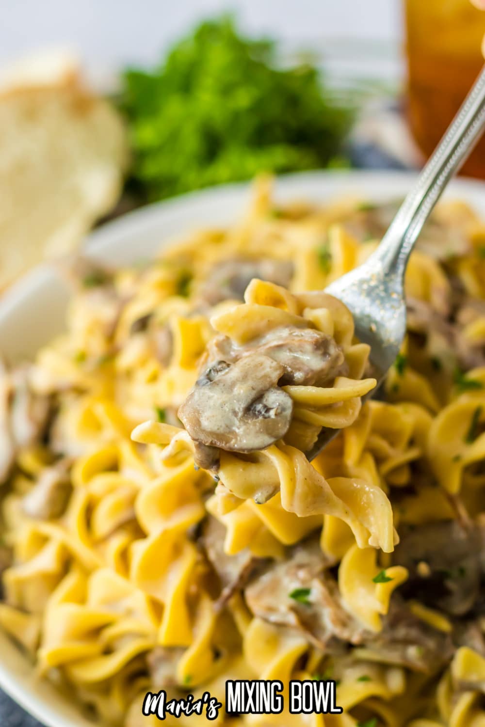 bite of mushrooms, beef, and egg noodles held on a fork over a white bowl filled with stroganoff