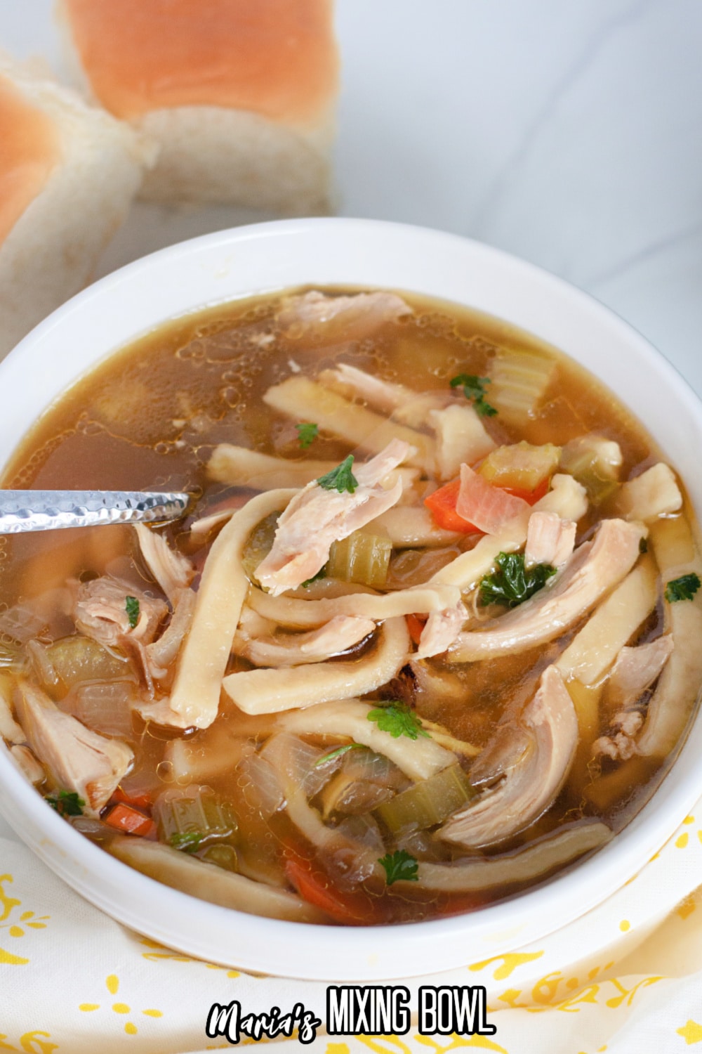 white bowl filled with chicken noodle soup with a sliver spoon with rolls in the background