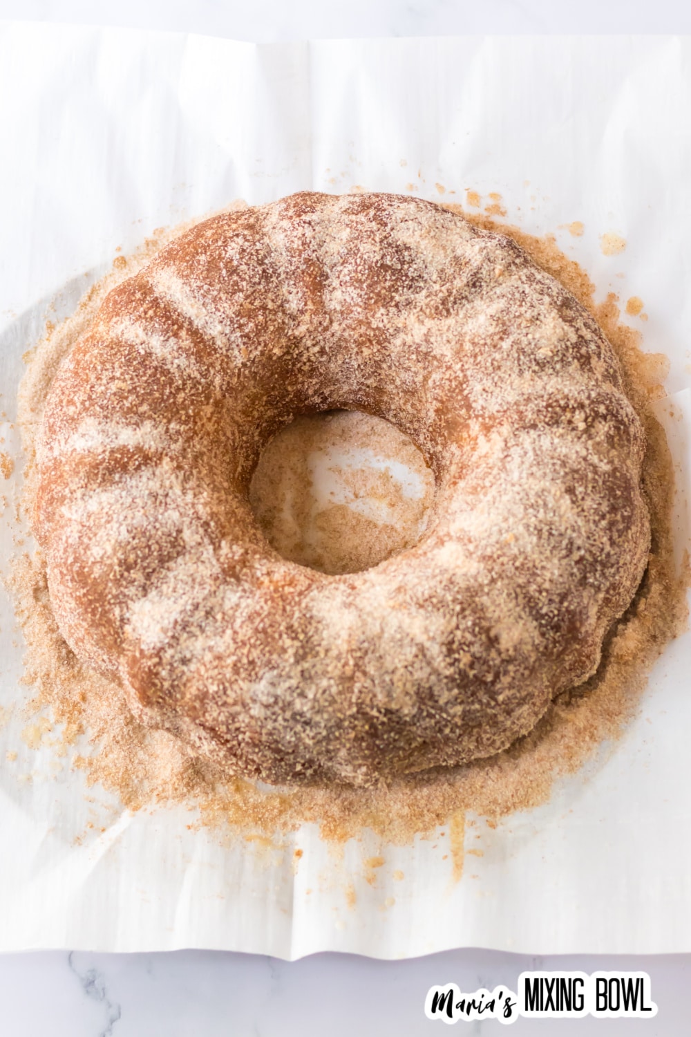 cinnamon sugar being sprinkled on top of a bundt cake on parchment paper.