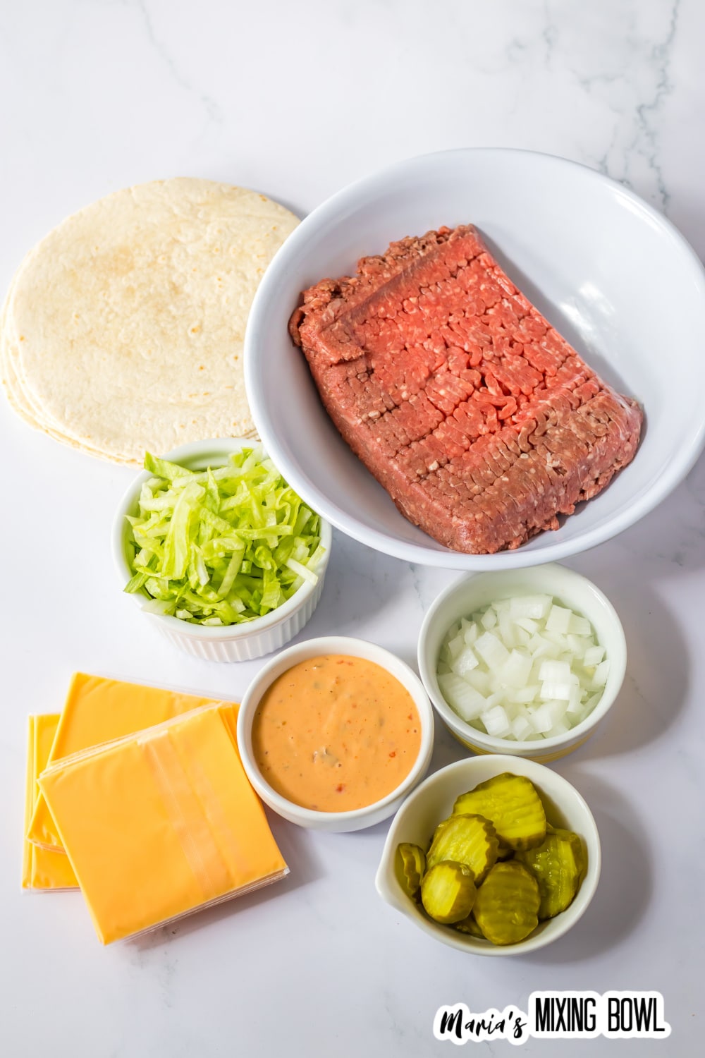 bowls of raw ground beef, shredded lettuce, sliced american cheese, big mac sauce, pickles, diced onions and tortillas.