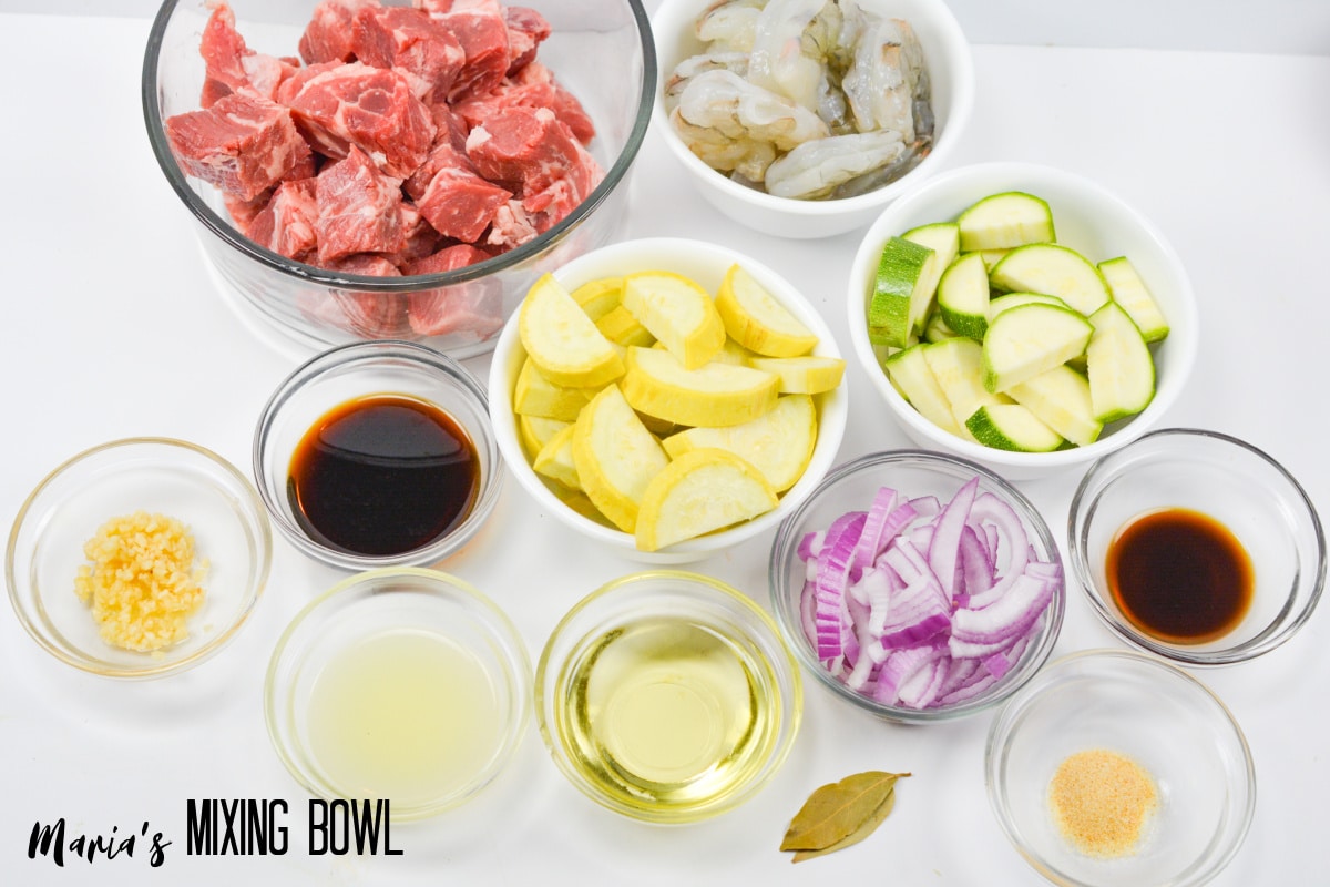 bowls of cut up steak, raw shrimp, yellow squash, zucchini squash, sliced red onions, garlic, bay leaves, oil, Worcestershire sauce, lemon juice, soy sauce, and garlic powder.