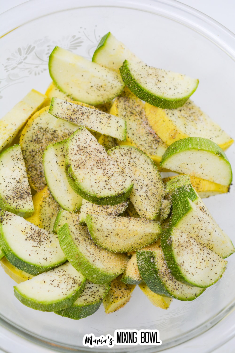 sliced raw zucchini and sliced yellow squash seasoning with salt and pepper in a glass bowl.
