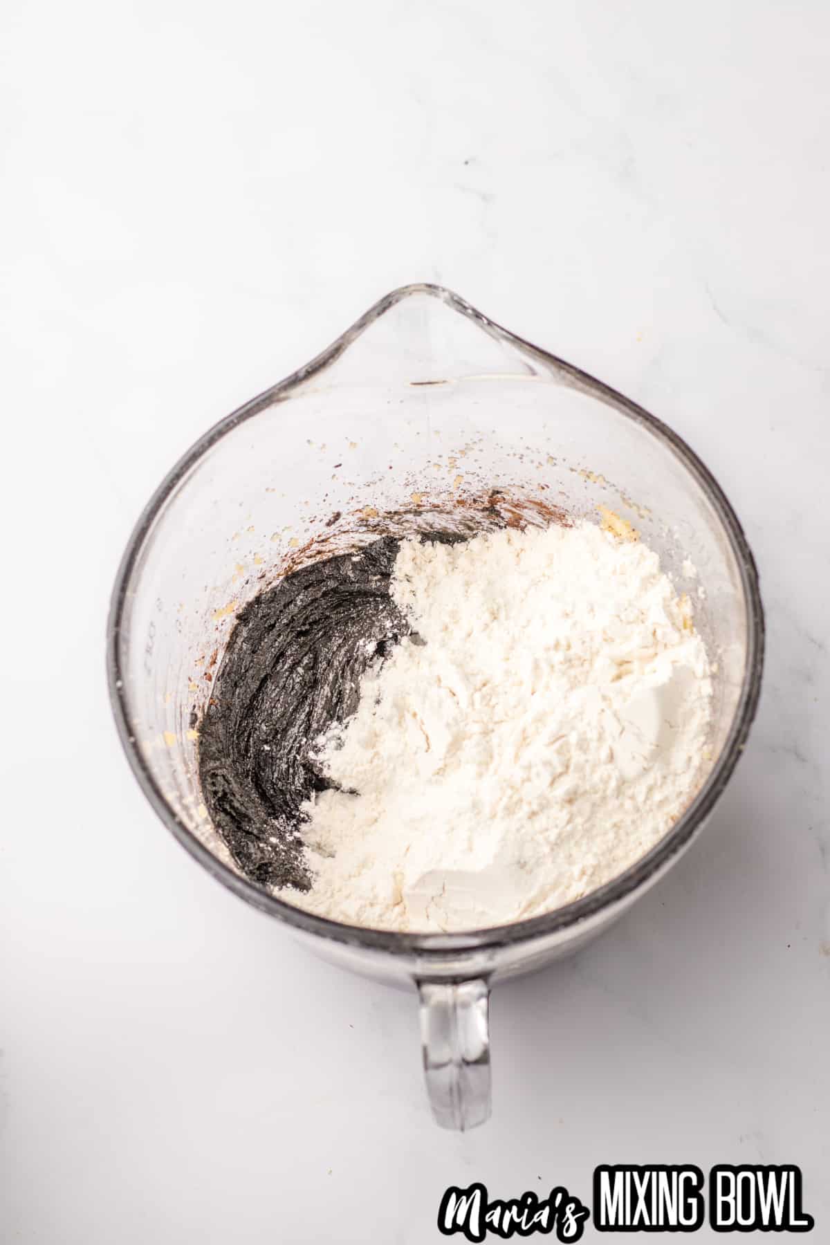 dry ingredients being added to black cookie dough in a glass mixing bowl.