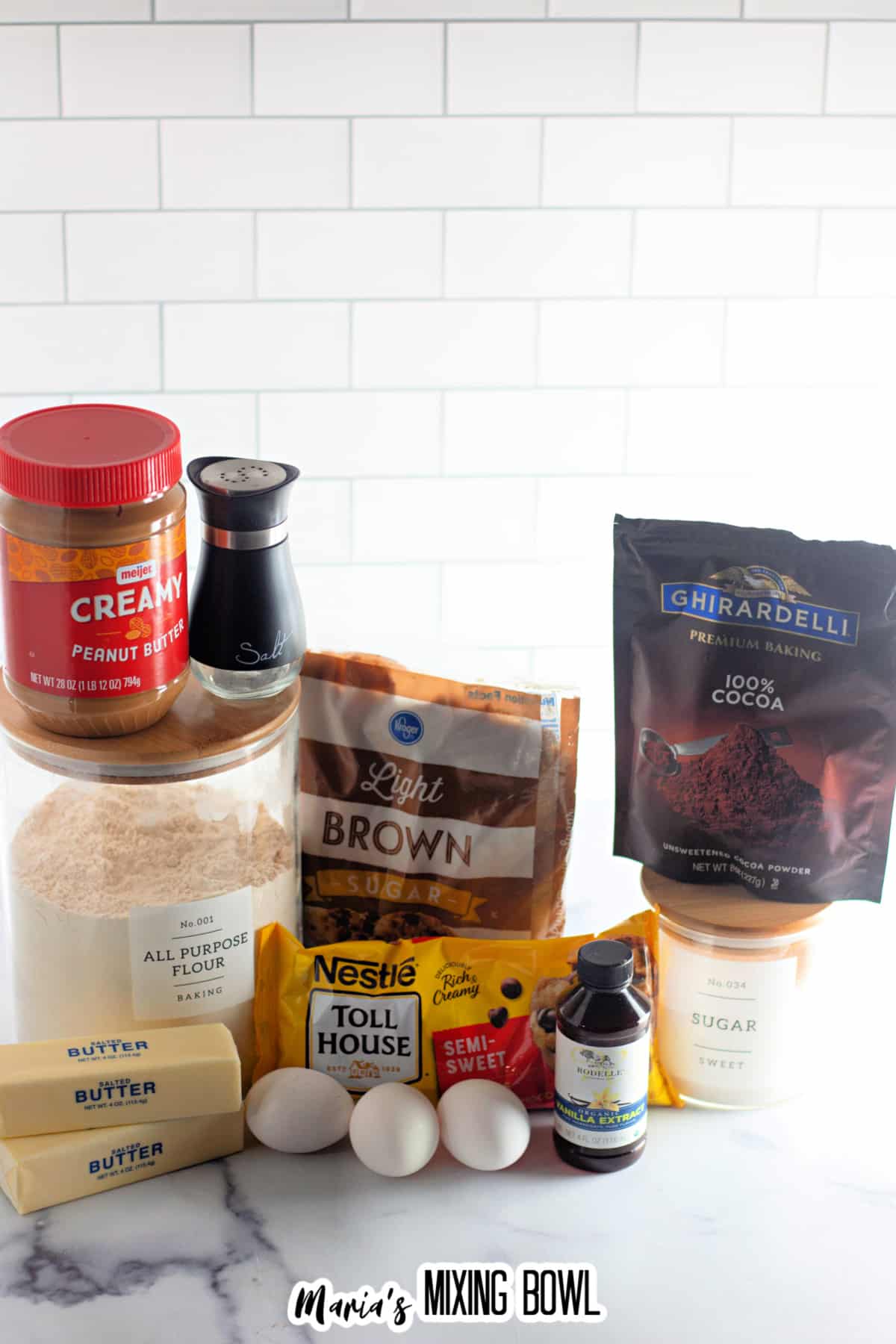 containers of peanut butter, salt, all purpose flour, butter, eggs, brown sugar, chocolate chips, vanilla, cocoa powder, and sugar on a counter.