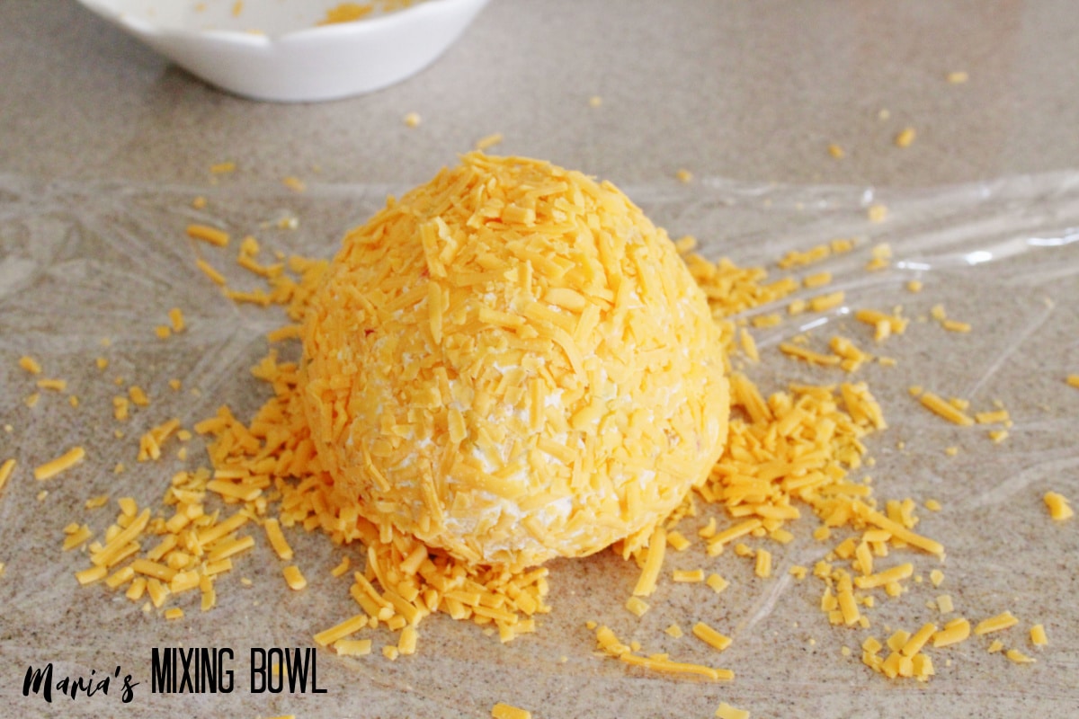 cheese ball being rolled in shredded cheddar cheese.