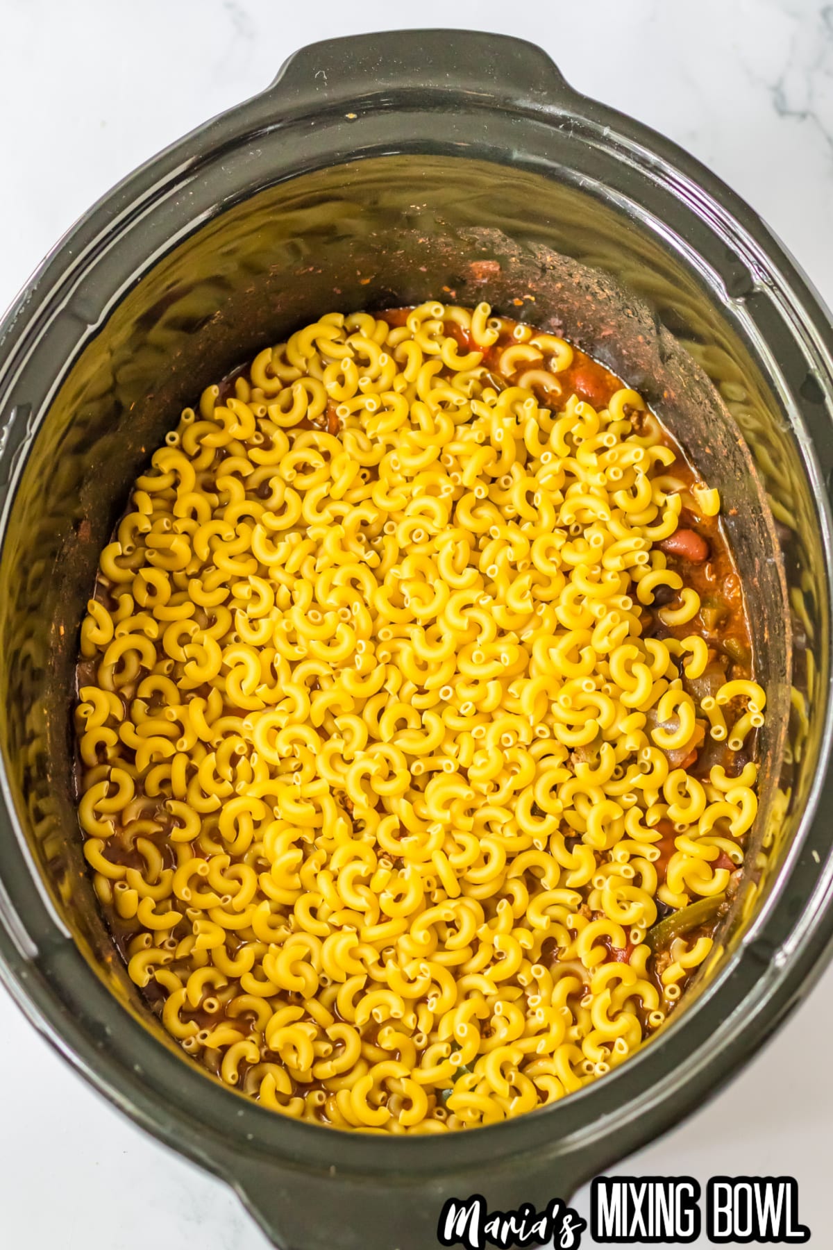 raw noodles being added to chili mac in a slow cooker.