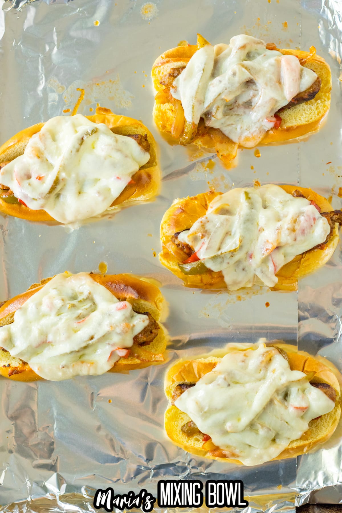 sausage and peppers on a hoagie roll with melted cheese on top on a foil lined baking sheet.