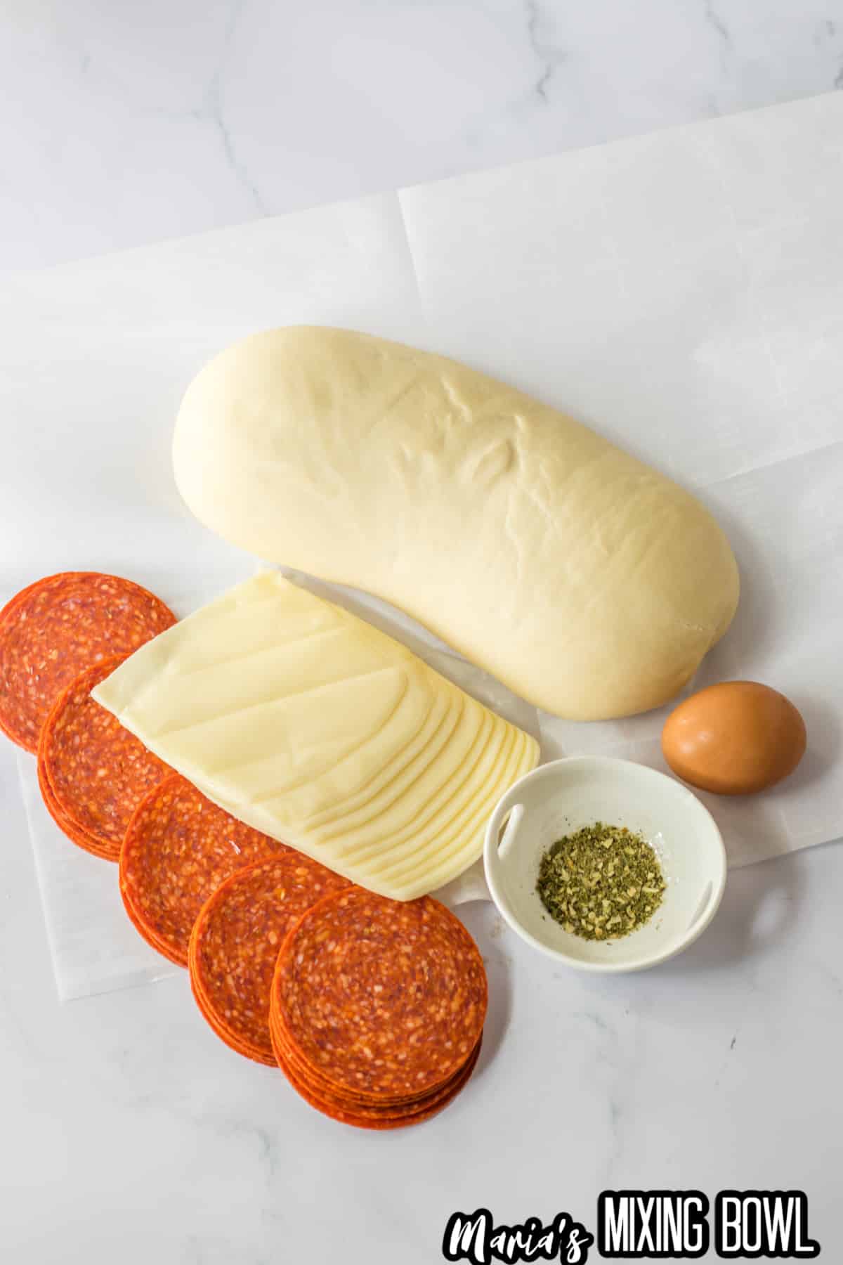 loaf of bread dough, slices of pepperoni, slices of mozzarella cheese, an egg, and a small bowl of italian seasoning.