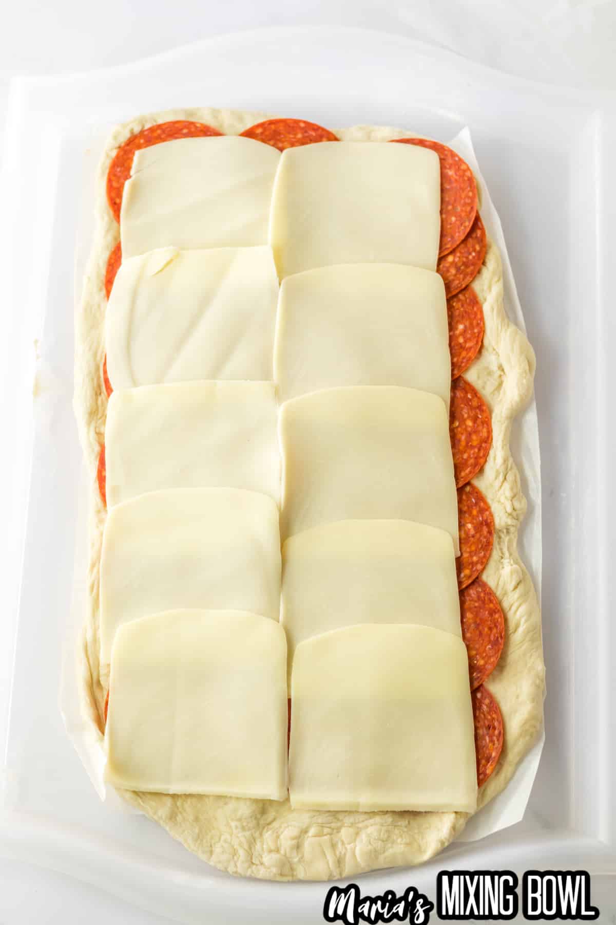 pepperoni slices and mozzarella slices being layered on rolled out raw bread dough.