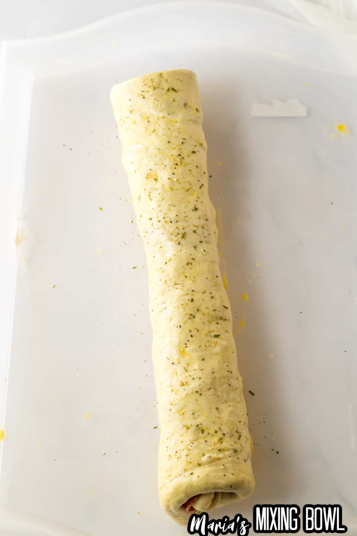 roll of stuffed pepperoni bread on a parchment baking sheet being brushed with egg wash and seasoned with italian seasoning.