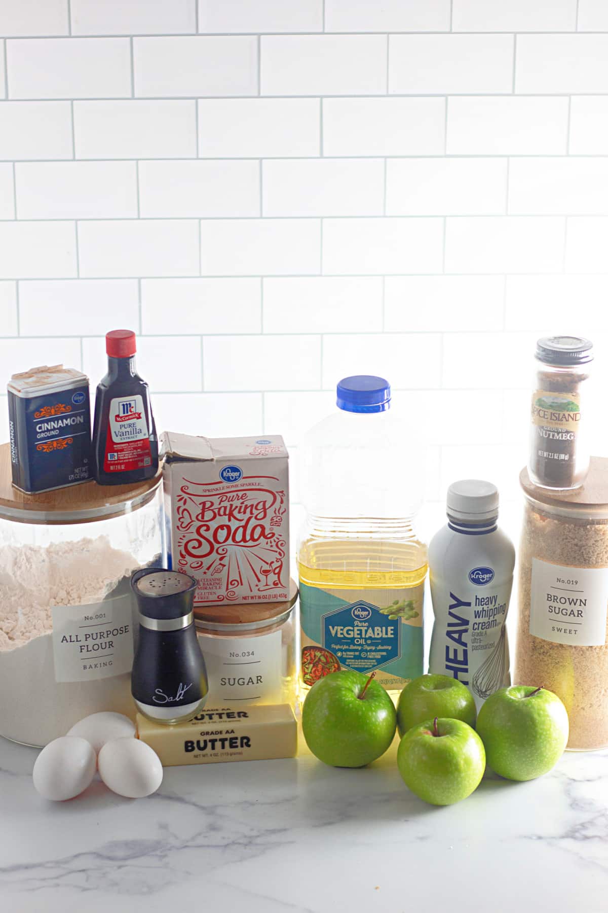 cinnamon, vanilla, flour, baking soda, sugar, salt, eggs, butter, apples, vegetable oil, heavy whipping cream, brown sugar and nutmeg on a counter.