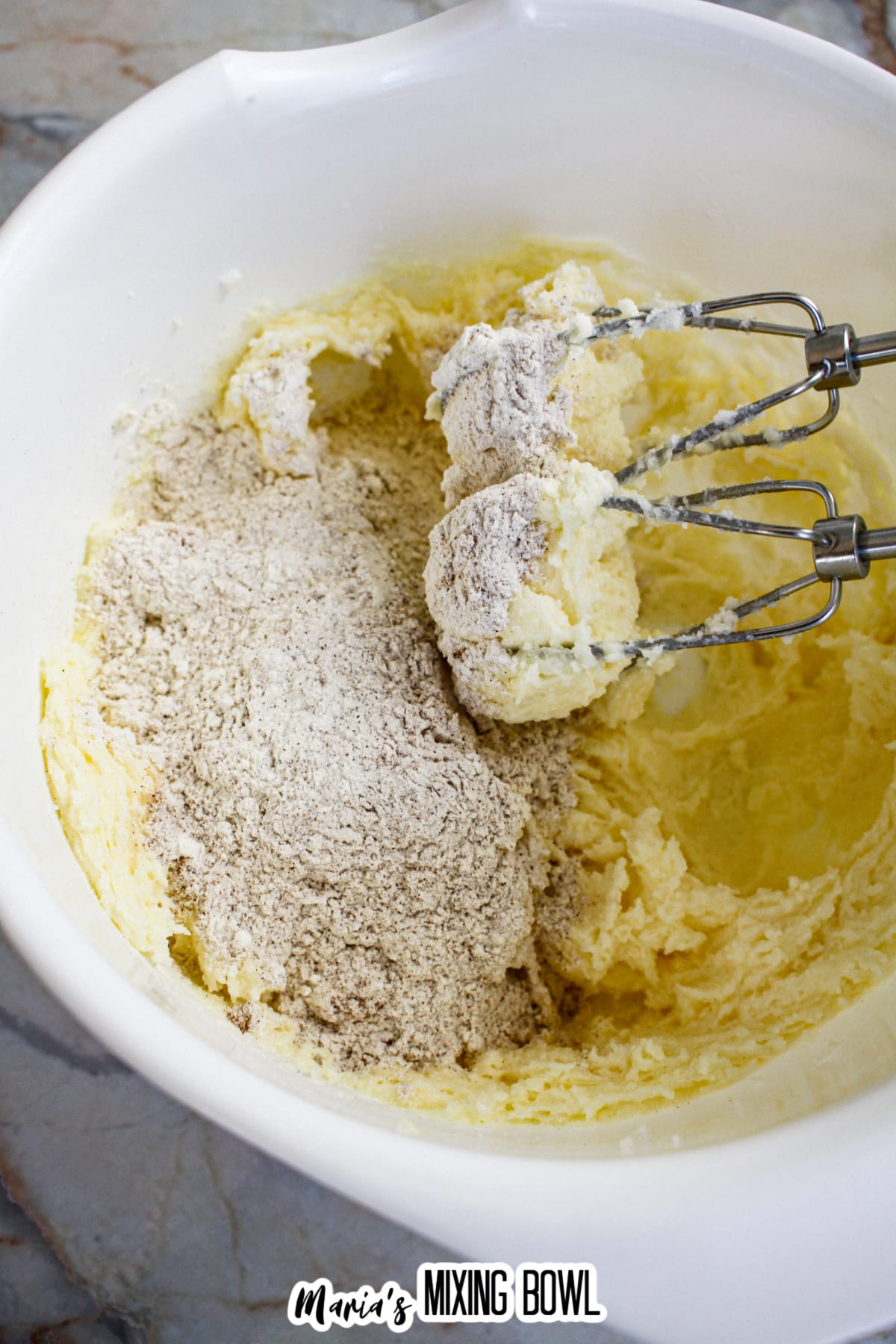 dry ingredients being added to the mixed white ingredients with a hand mixer in a mixing bowl.