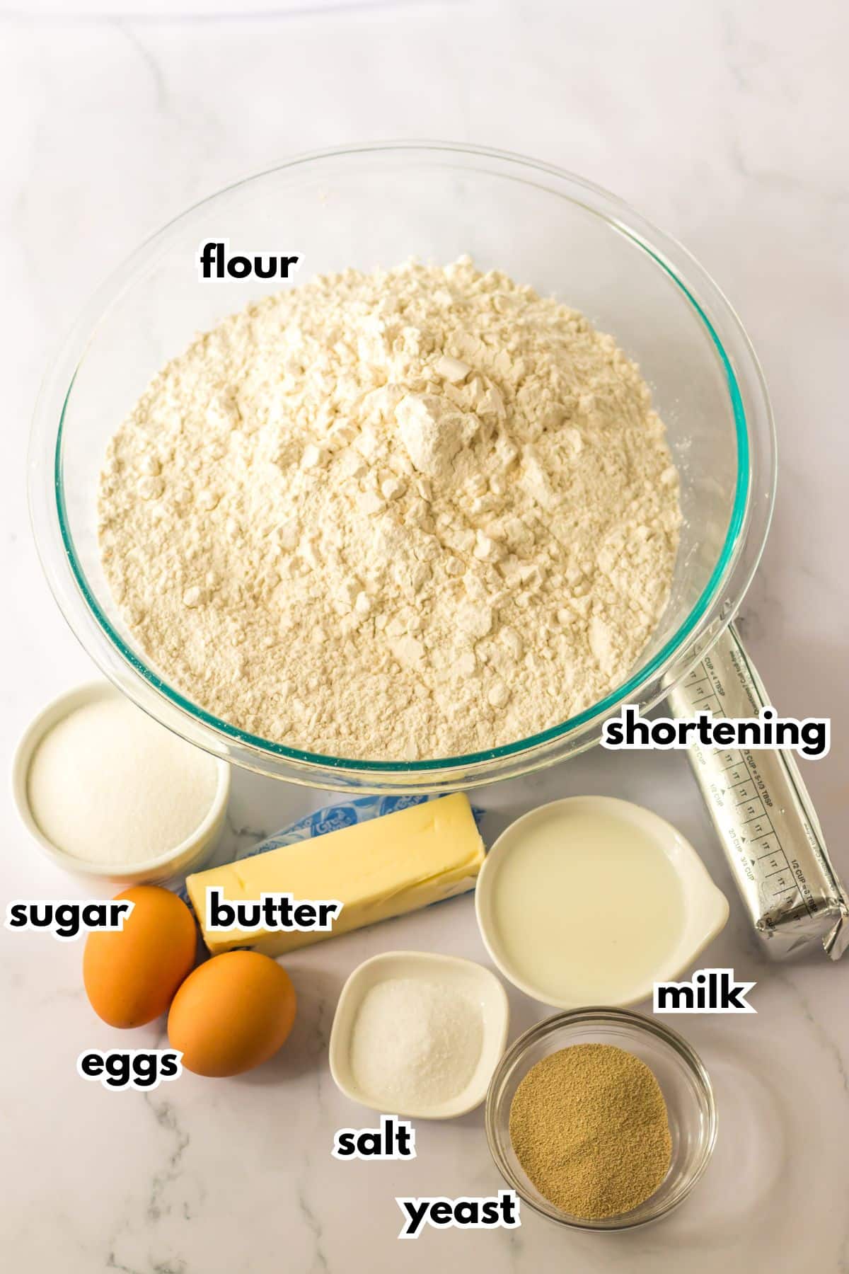 bowls of flour, sugar, butter, eggs, salt, yeast, milk, and shortening on a counter.