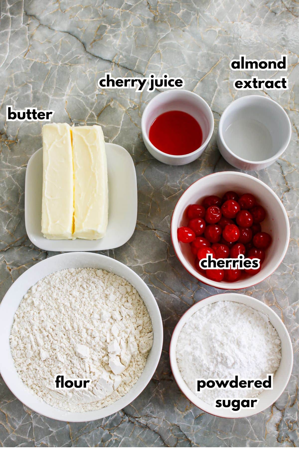 bowls of butter, cherry juice, almond extract, flour, cherries, and powdered sugar on a counter.