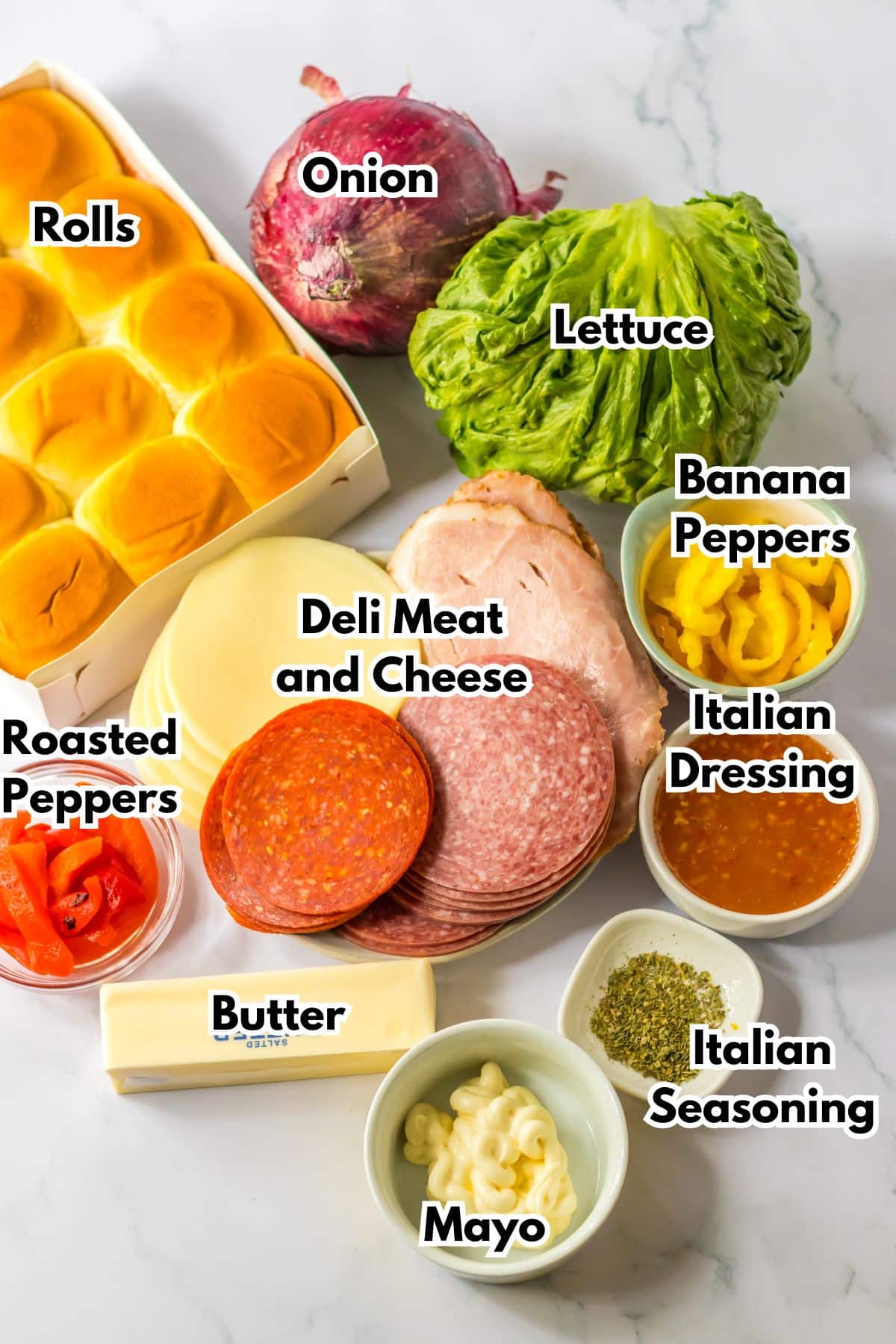 rolls, onion, lettuce, banana peppers, roasted peppers, deli meat and cheese, italian dressing, butter, mayo, italian seasoning on a counter.