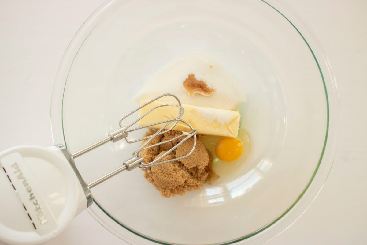 butter, brown sugar, egg, and vanilla being mixed with a hand mixer in a glass mixing bowl.