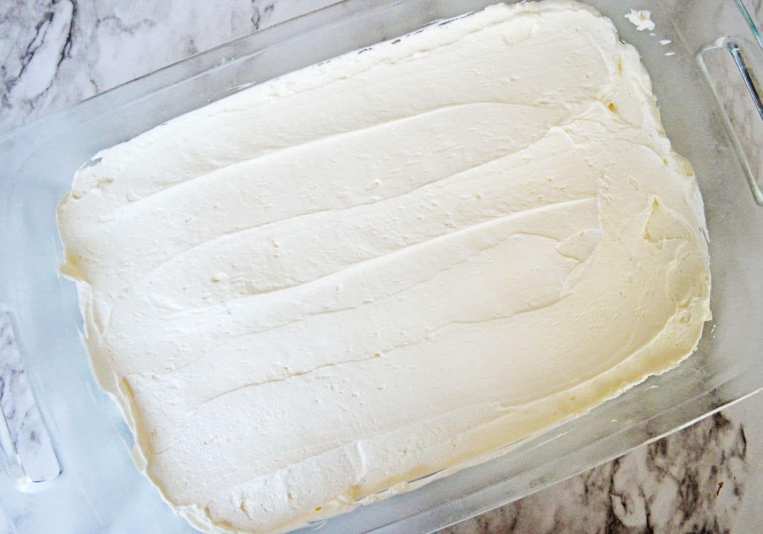 cream cheese layer being spread on top of an golden oreo cookie crust in a glass baking dish.