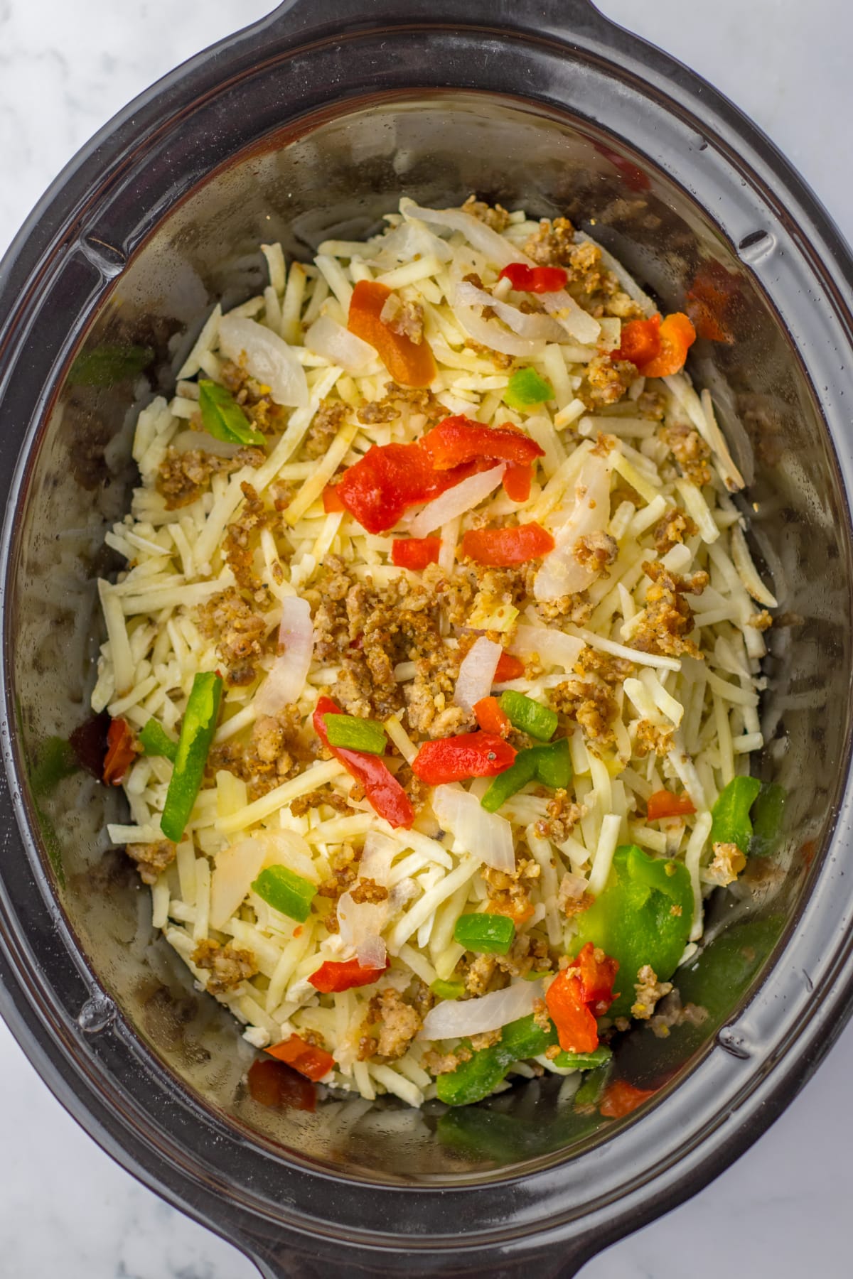 hashbrowns, peppers, onions, and cooked sausage being layered in a slow cooker.