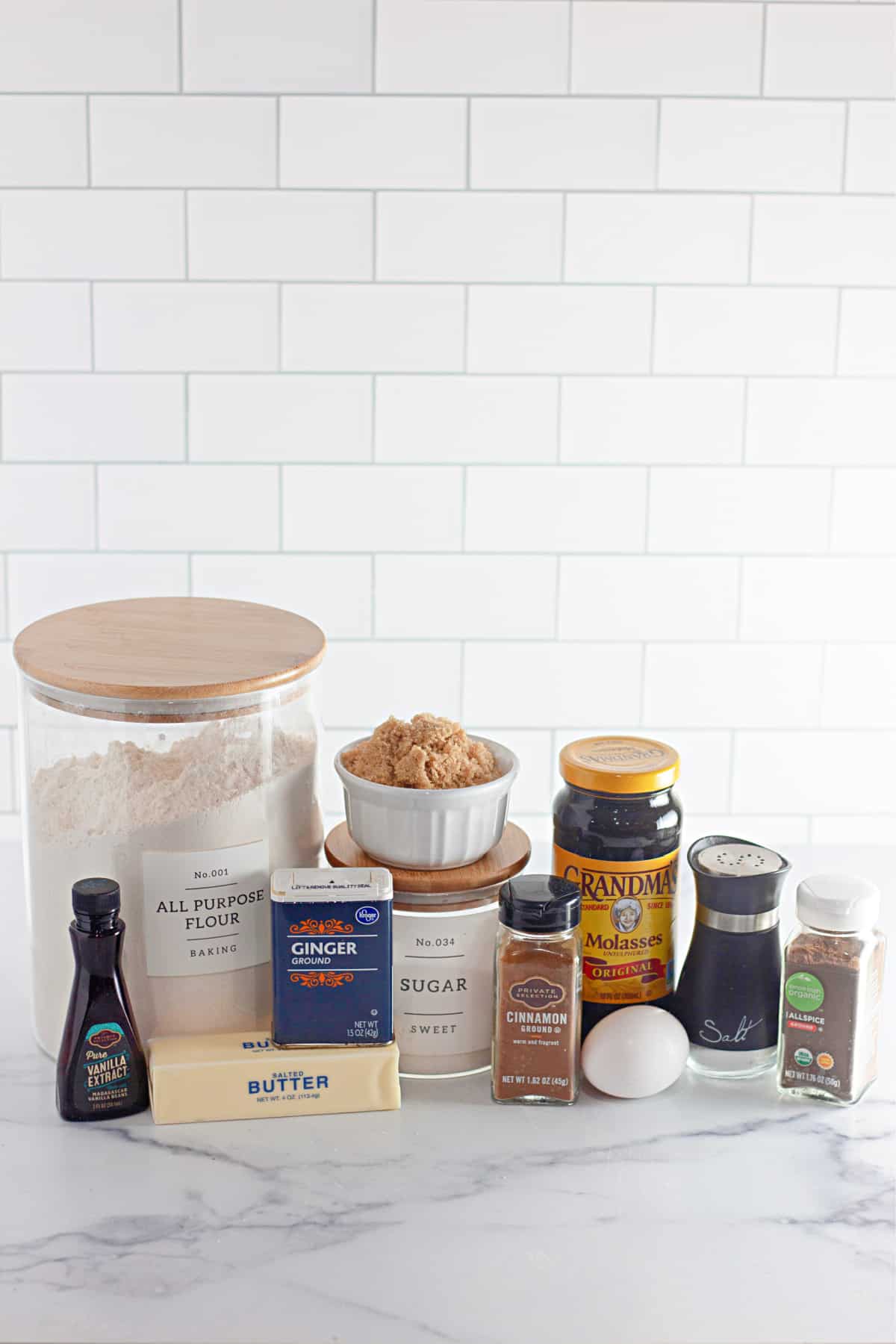 containers of flour, vanilla, ginger, butter, brown sugar, sugar, cinnamon, eggs, molasses, salt, and allspice on a counter.