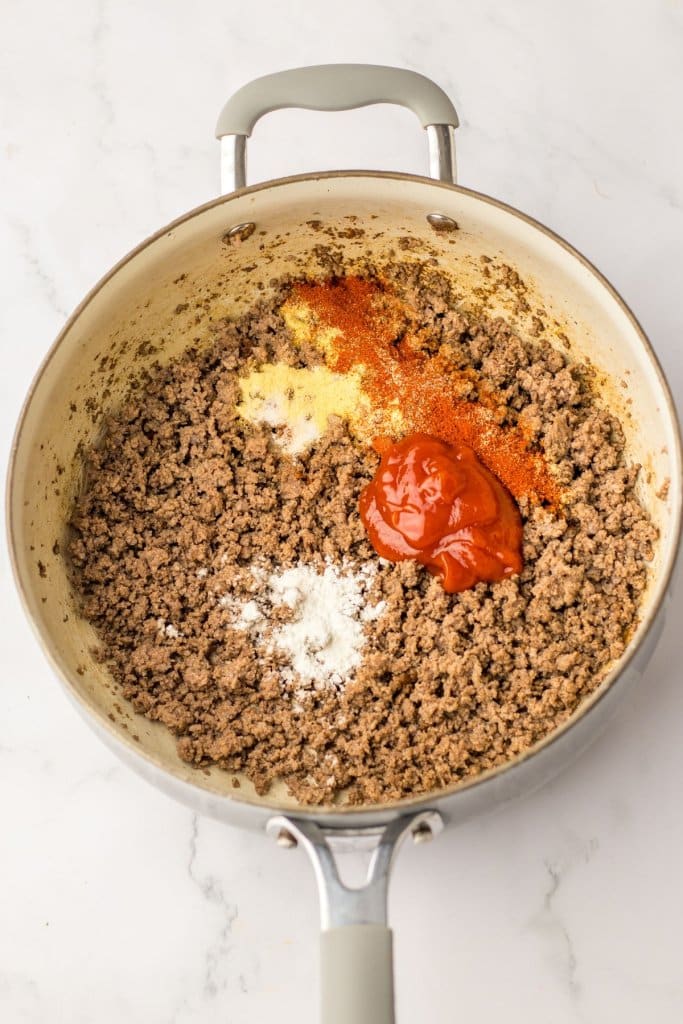 ketchup, and seasonings being added to cooked ground beef in a skillet.
