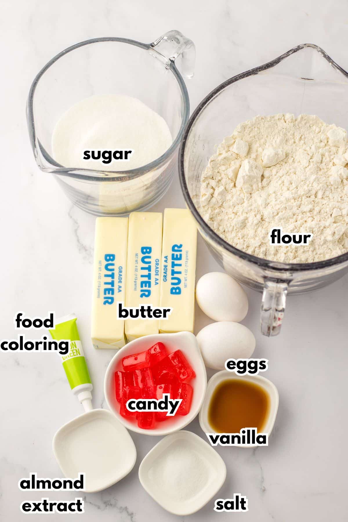 bowls of sugar, flour, butter, food coloring, eggs, candy, almond extract, salt, and vanilla extract on a counter.