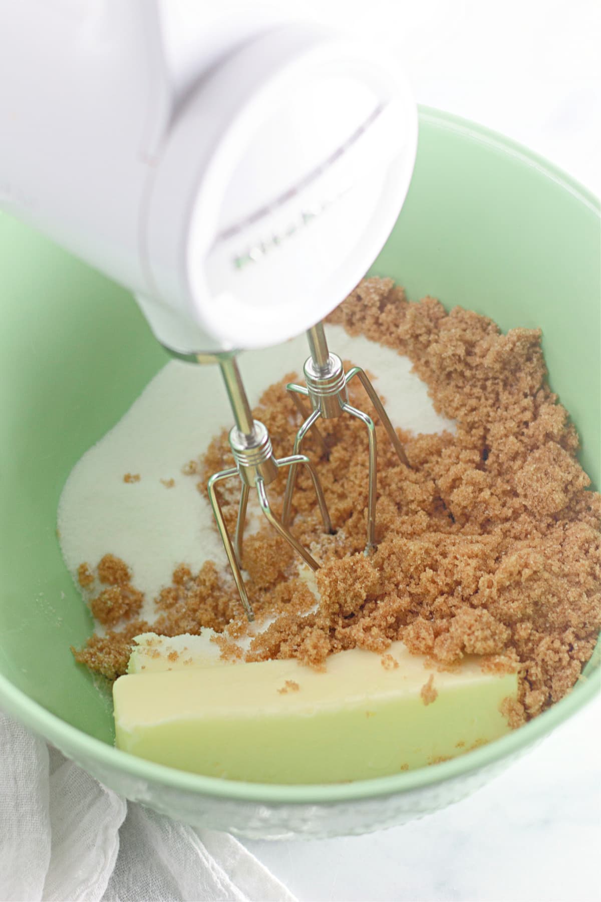butter, sugar, and brown sugar being mixed with a hand mixer in a bowl.