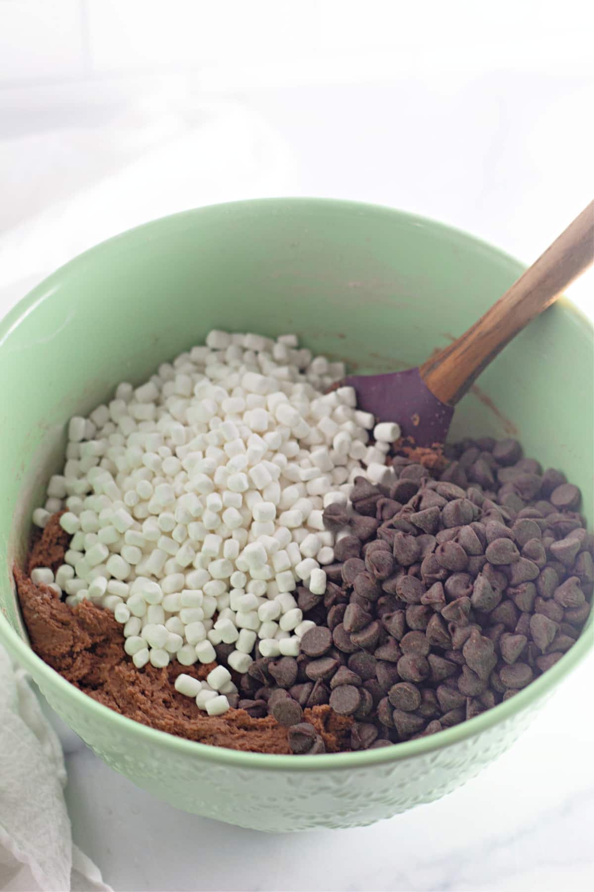 mini marshmallows and chocolate chips being mixed into cookie dough in a mixing bowl.