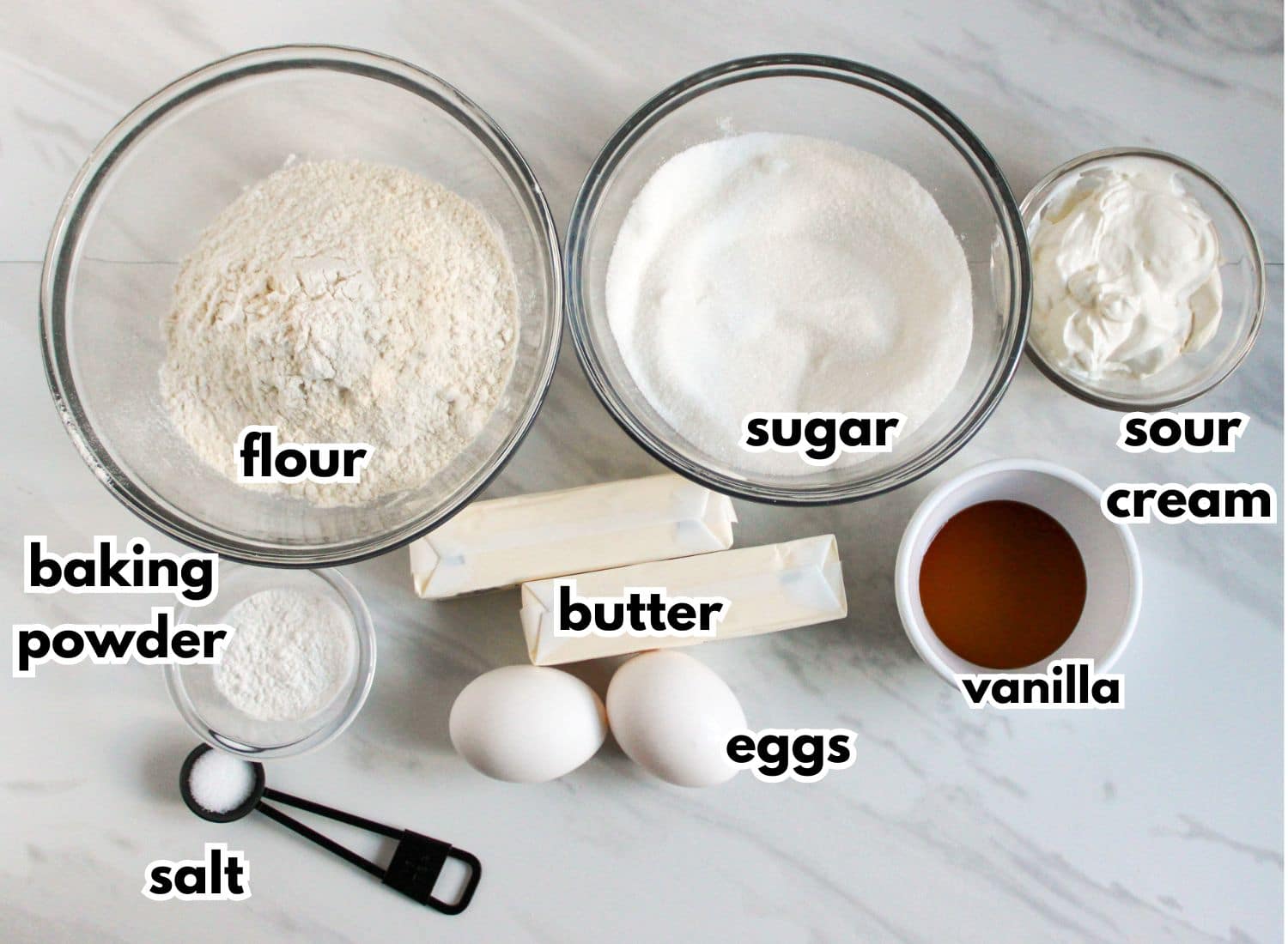 bowls of flour, sugar, sour cream, baking powder, butter, eggs, vanilla extract, and salt on a counter.