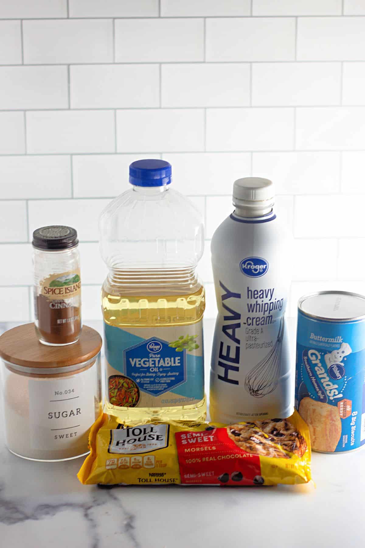 containers of cinnamon, sugar, vegetable oil, heavy cream, can of biscuits, and chocolate chips on a counter.