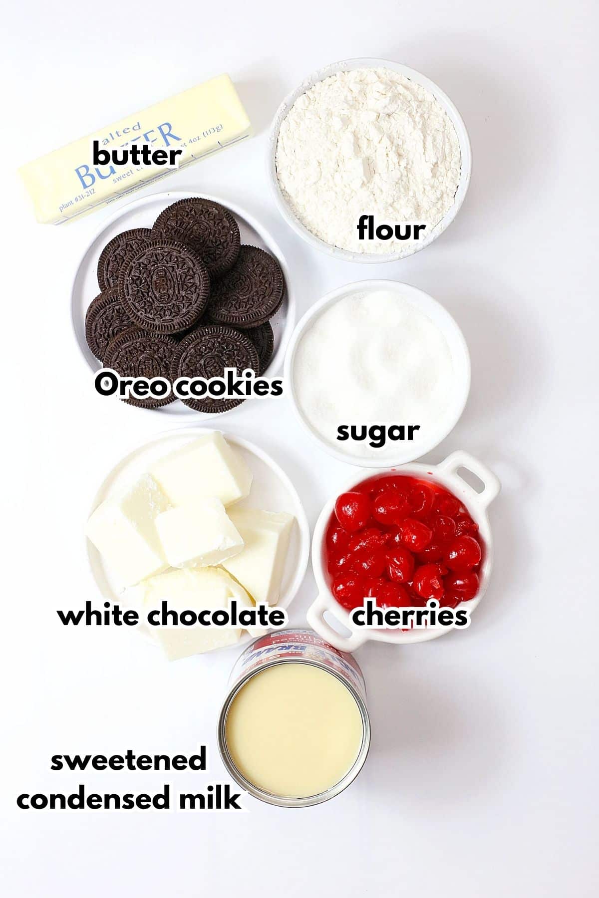 bowls of butter, flour, Oreo cookies, white chocolate, sweetened condensed milk, sugar, and cherries on a counter.