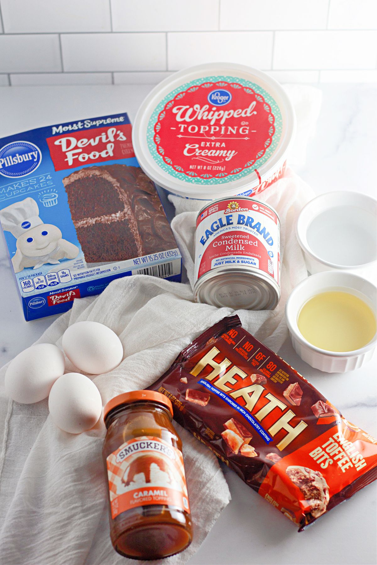 box of cake mix, can of condensed milk, jar of caramel sauce, container of whipped toppings, eggs, oil and water on a counter.