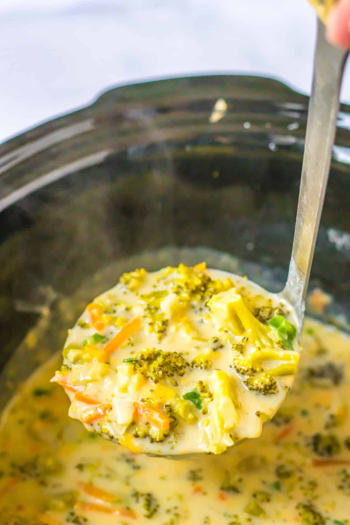 ladle full of panera broccoli cheddar soup held in the air above a slow cooker.