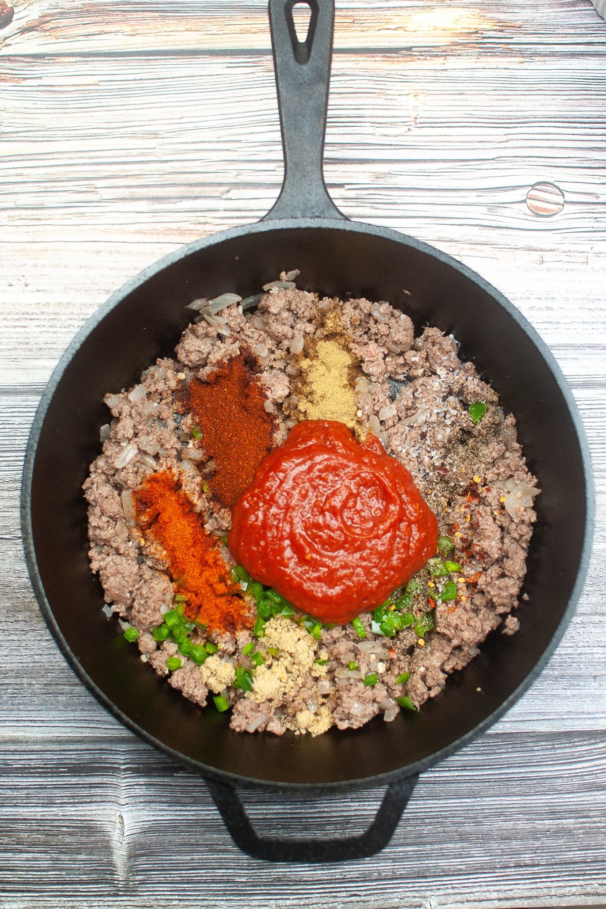 diced jalapeno, seasonings, and tomatoes being added to cooked ground beef in a cast iron skillet.