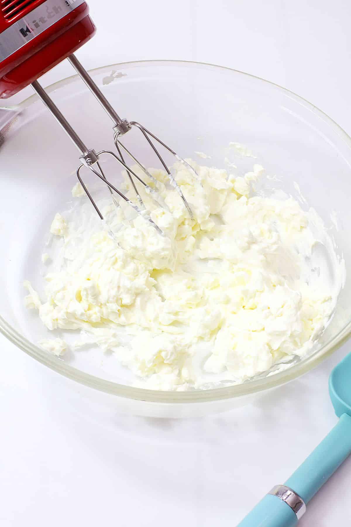 cream cheese and powdered sugar being mixed with a hand mixer in a glass bowl.