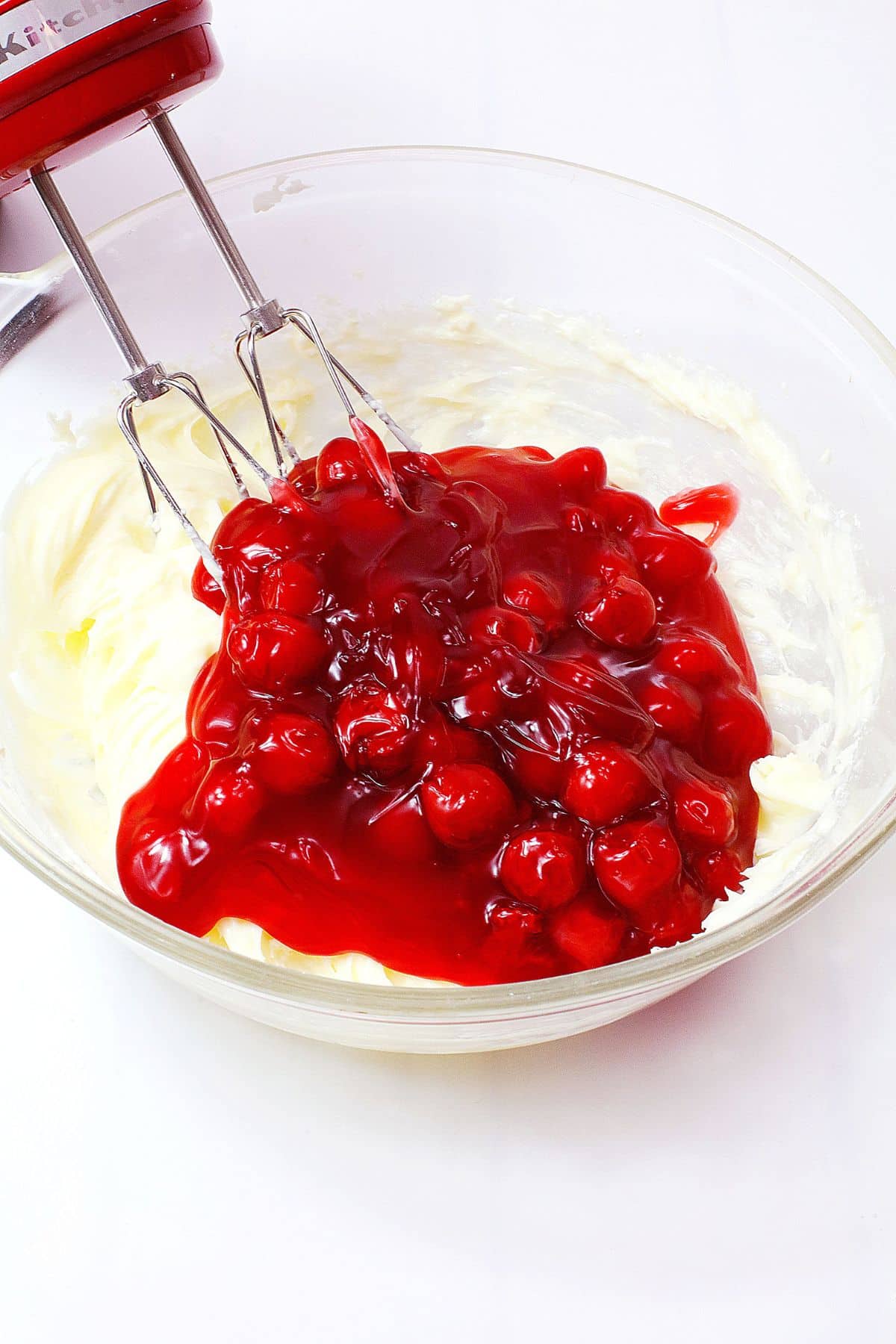 cherry pie filling being added to cream cheese in a glass mixing bowl.