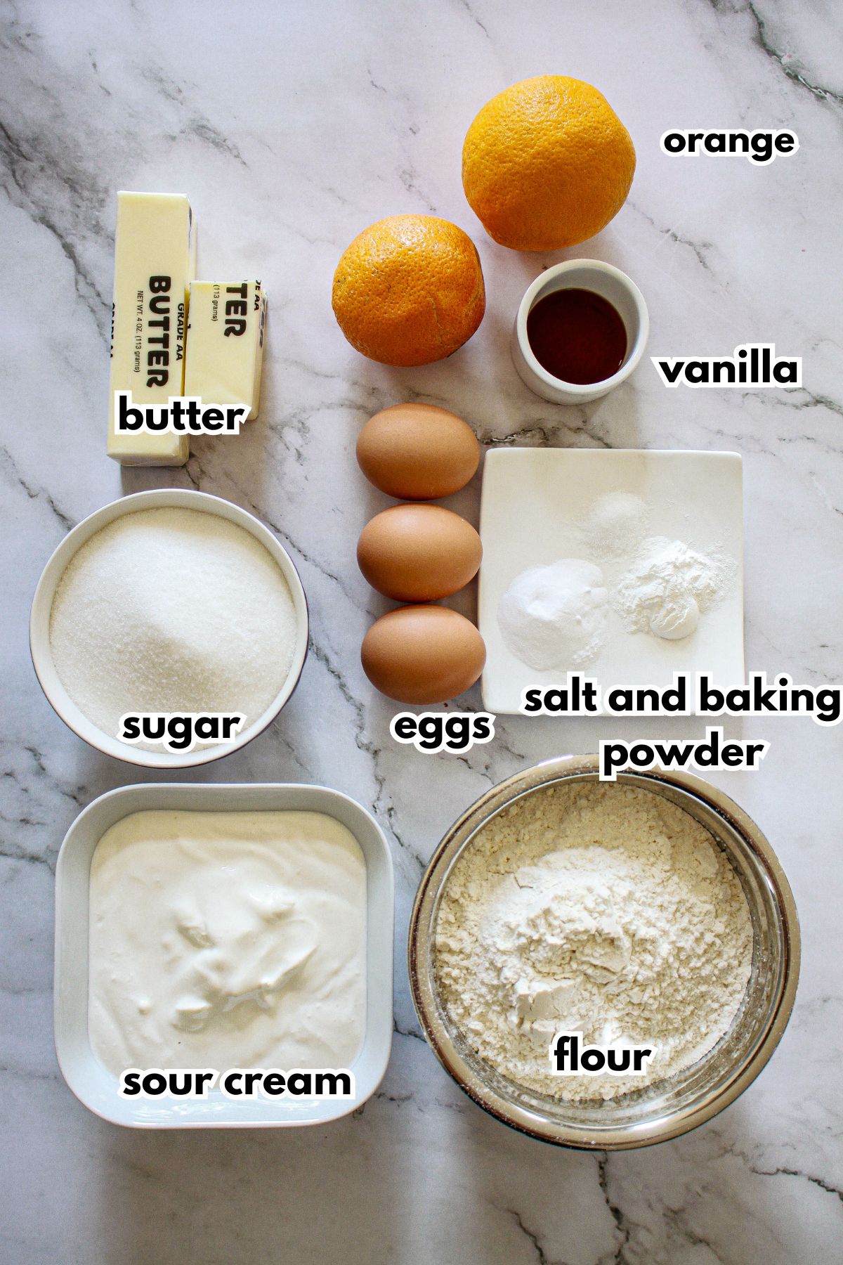 butter, oranges, vanilla, eggs, salt and baking powder, sour cream, and flour on a marble counter.