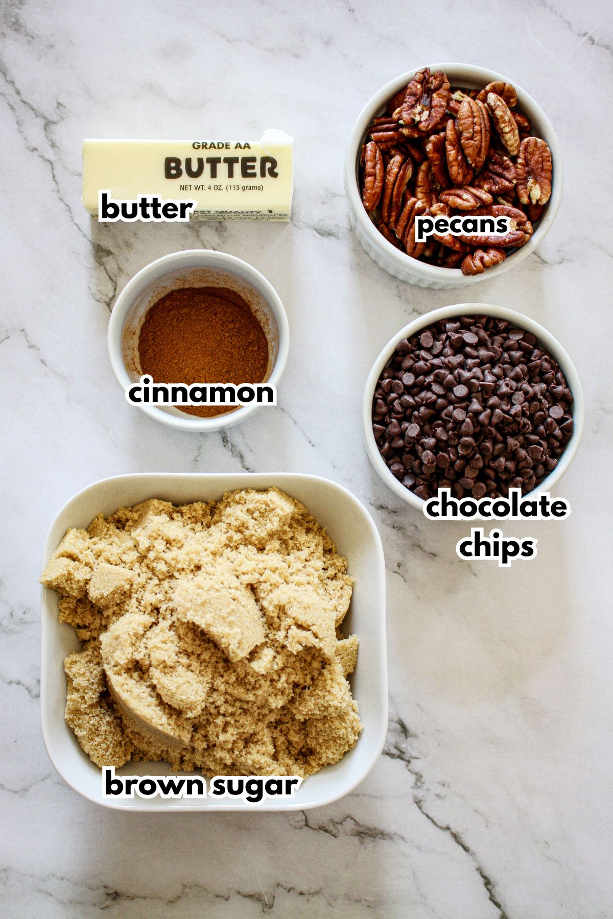 bowls of butter, cinnamon, brown sugar, pecans, and chocolate chips on a marble counter.
