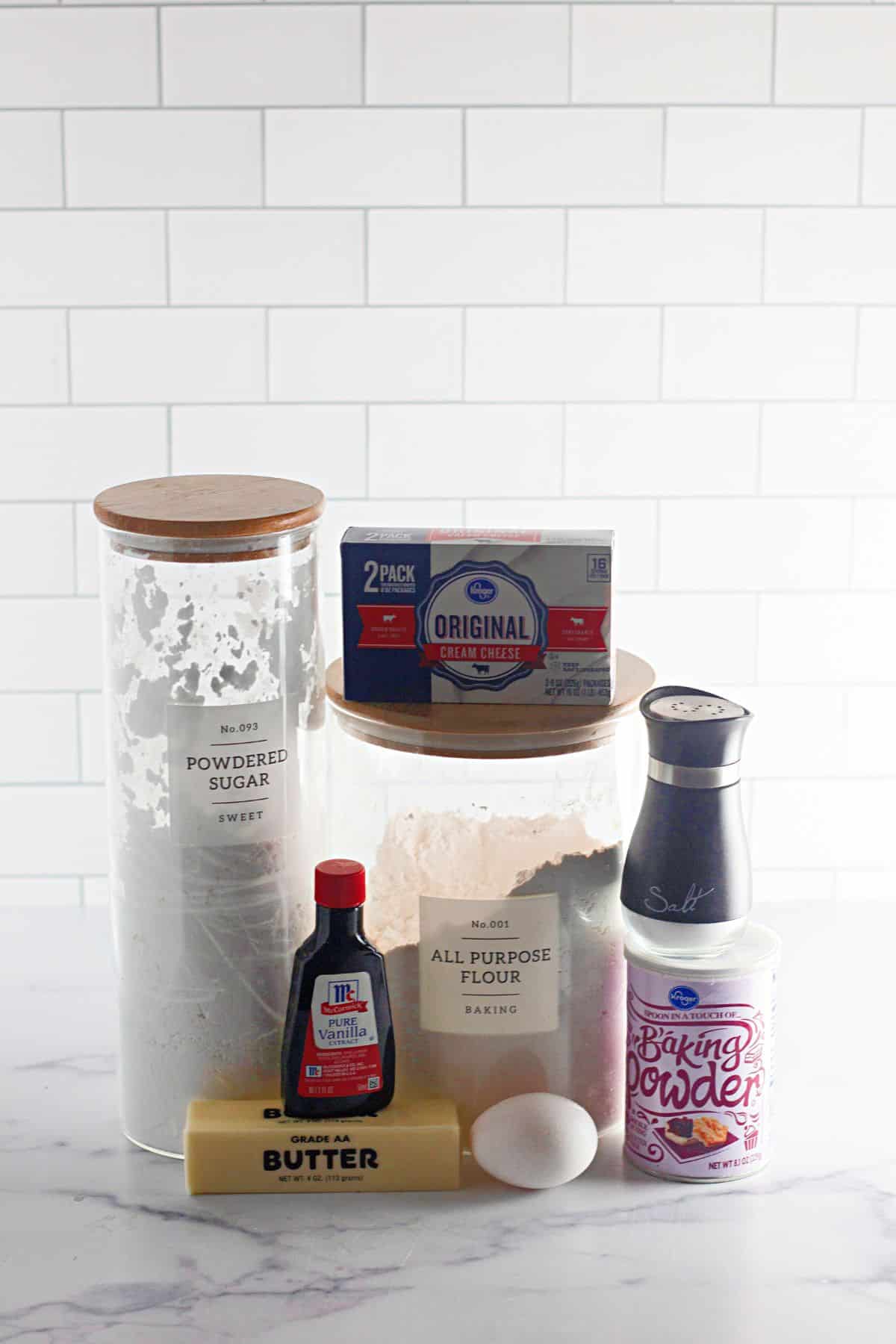 containers of powdered sugar, flour, cream cheese, vanilla, butter, egg, salt and baking powder on a counter.