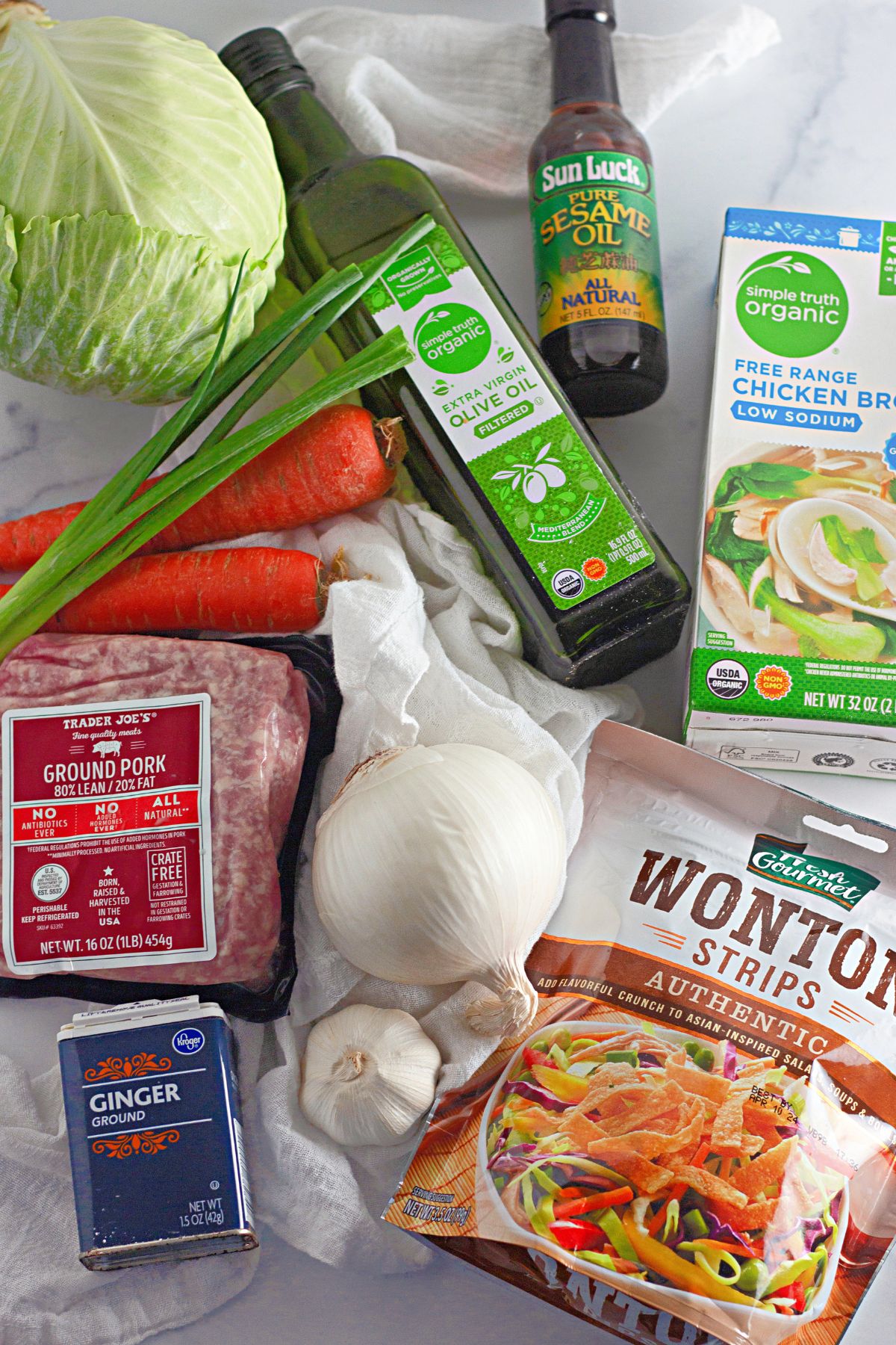 head of cabbage, two carrots, green onions, olive oil, sesame oil, chicken broth, ground pork, ginger, onion, garlic and wonton strips on a counter.