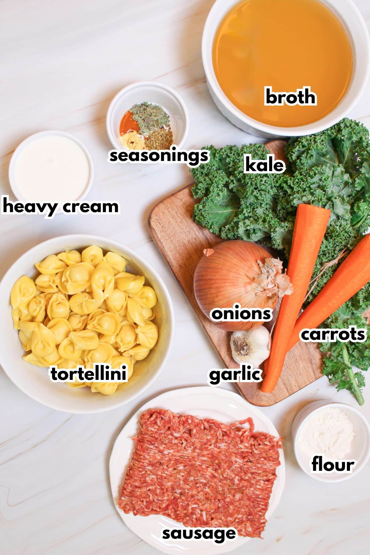 bowls of broth, seasonings, heavy cream, tortellini, sausage, onion, garlic, carrots, kale, and flour on a counter.