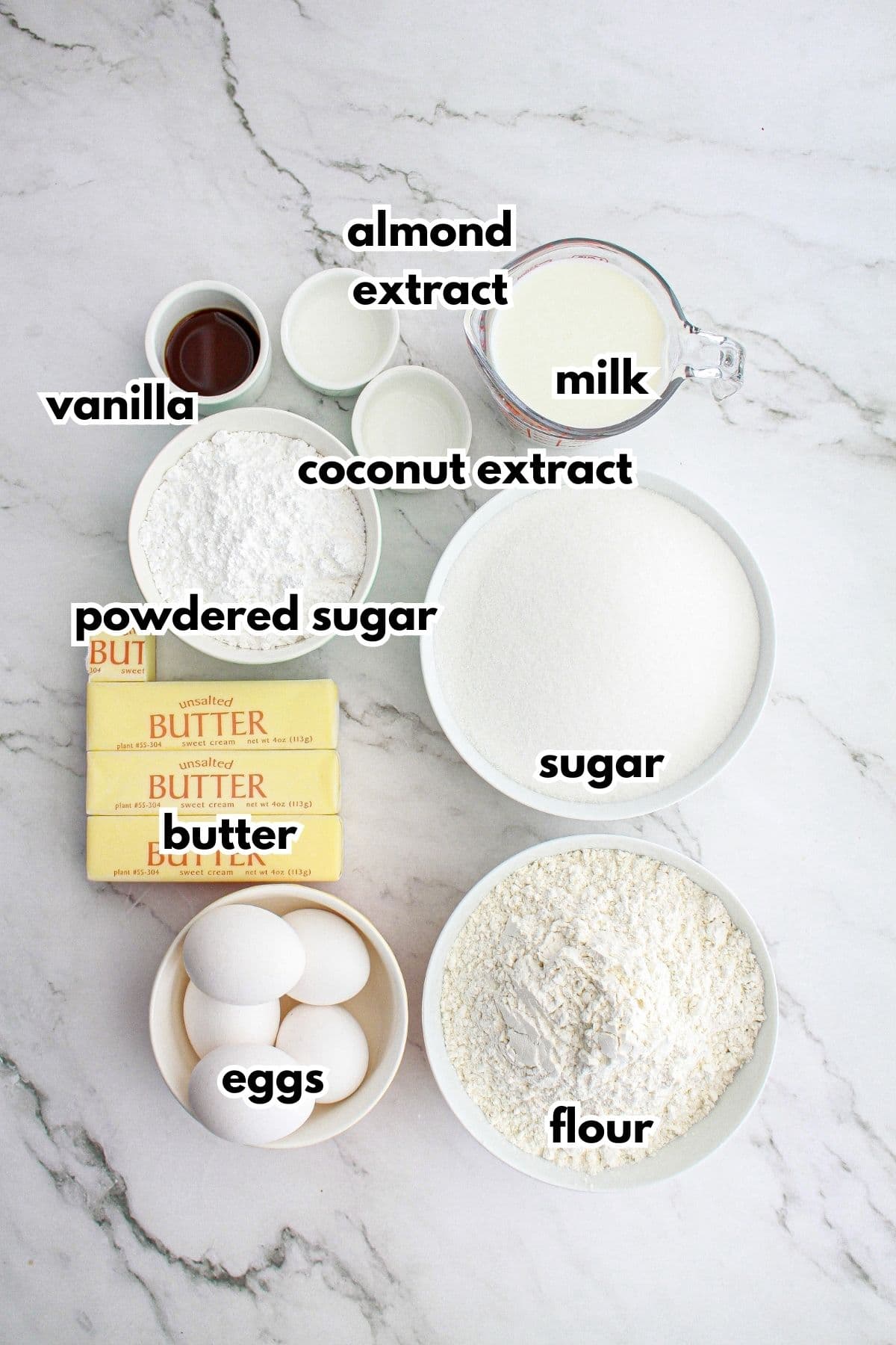 bowls of vanilla, almond extract, coconut extract, powdered sugar, butter, eggs, flour, and sugar on a counter.