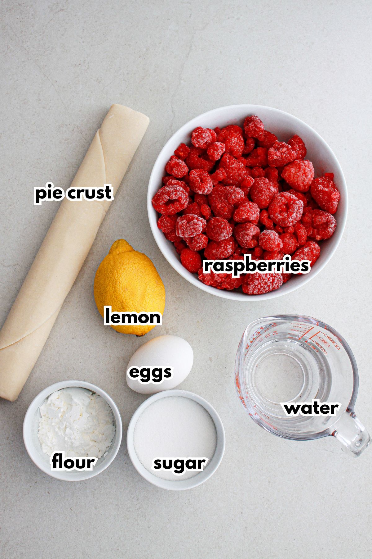 bowls of raspberries, pie crust, lemon, egg, flour, sugar, and water on a counter.