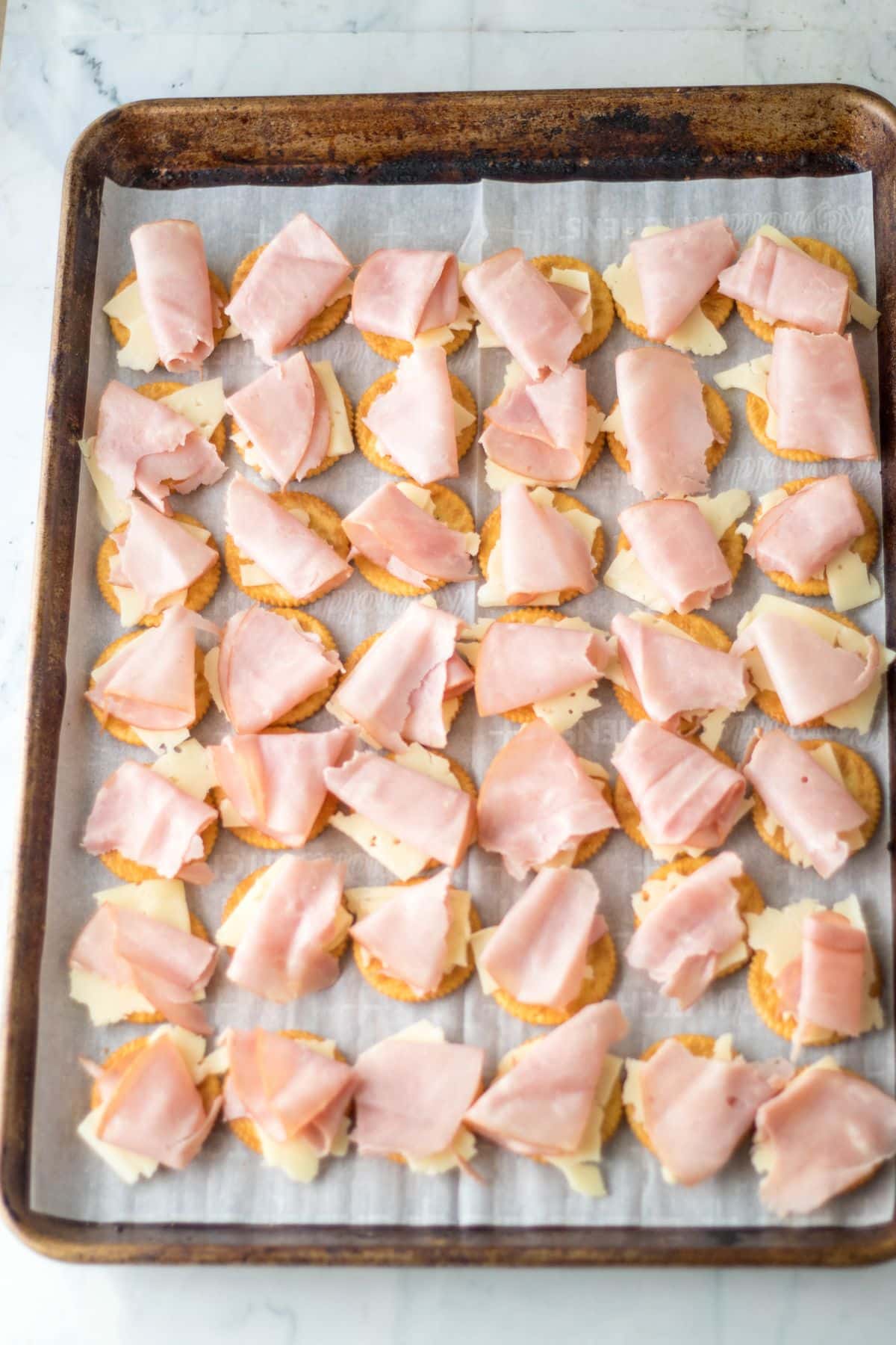pieces of ham and sliced cheese being placed on top of crackers on a parchment lined baking sheet.