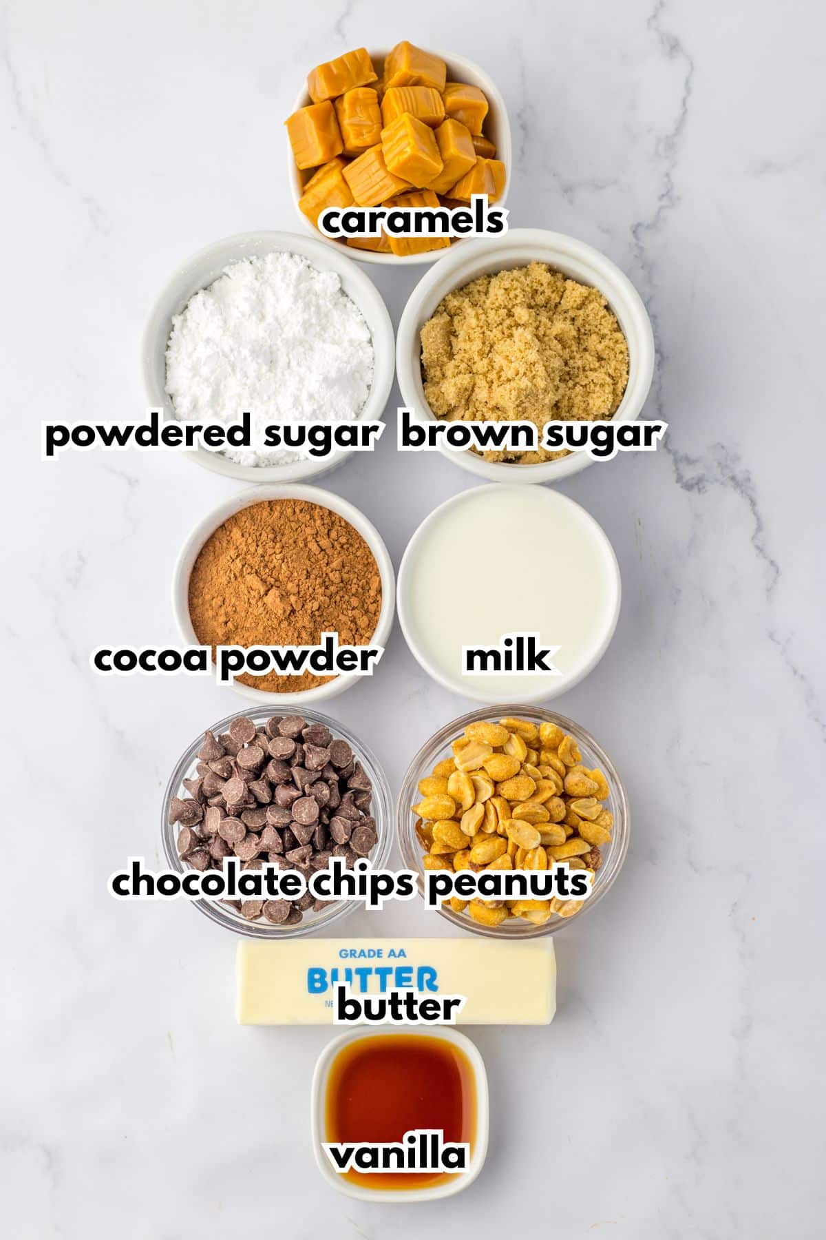 bowls of caramels, powdered sugar, brown sugar, cocoa powder, milk, chocolate chips, peanuts, butter, and vanilla on a counter.